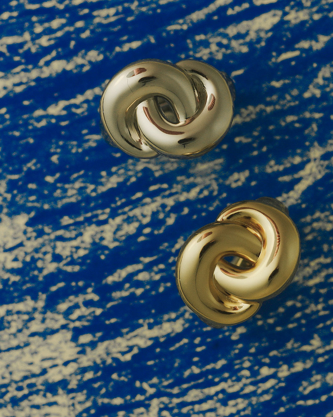The Luv Aj Solstice Knot Studs in Gold on blue background