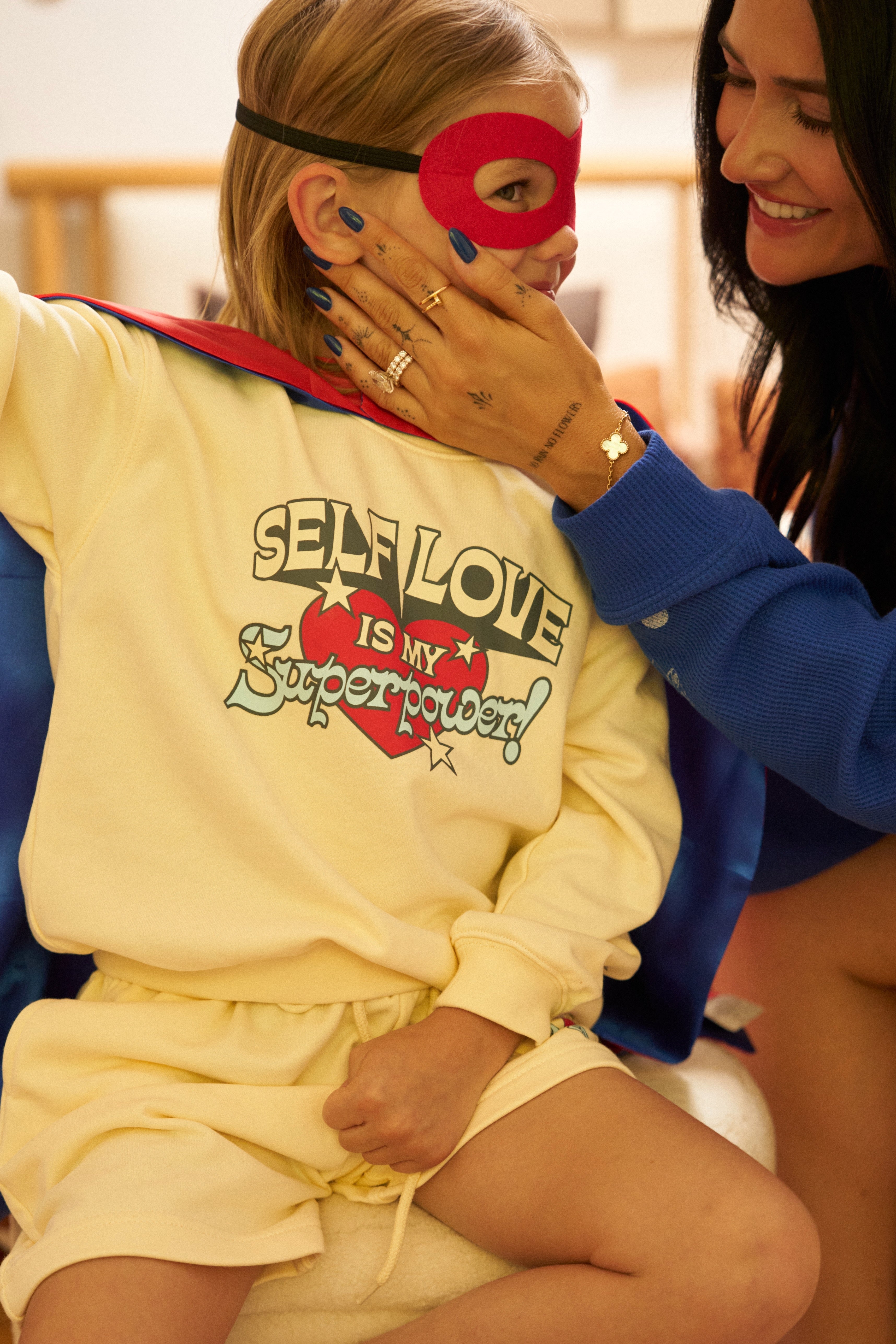 Child in superhero costume with 'Self Love is My Superpower' shirt, wearing a red mask, with adult beside them.