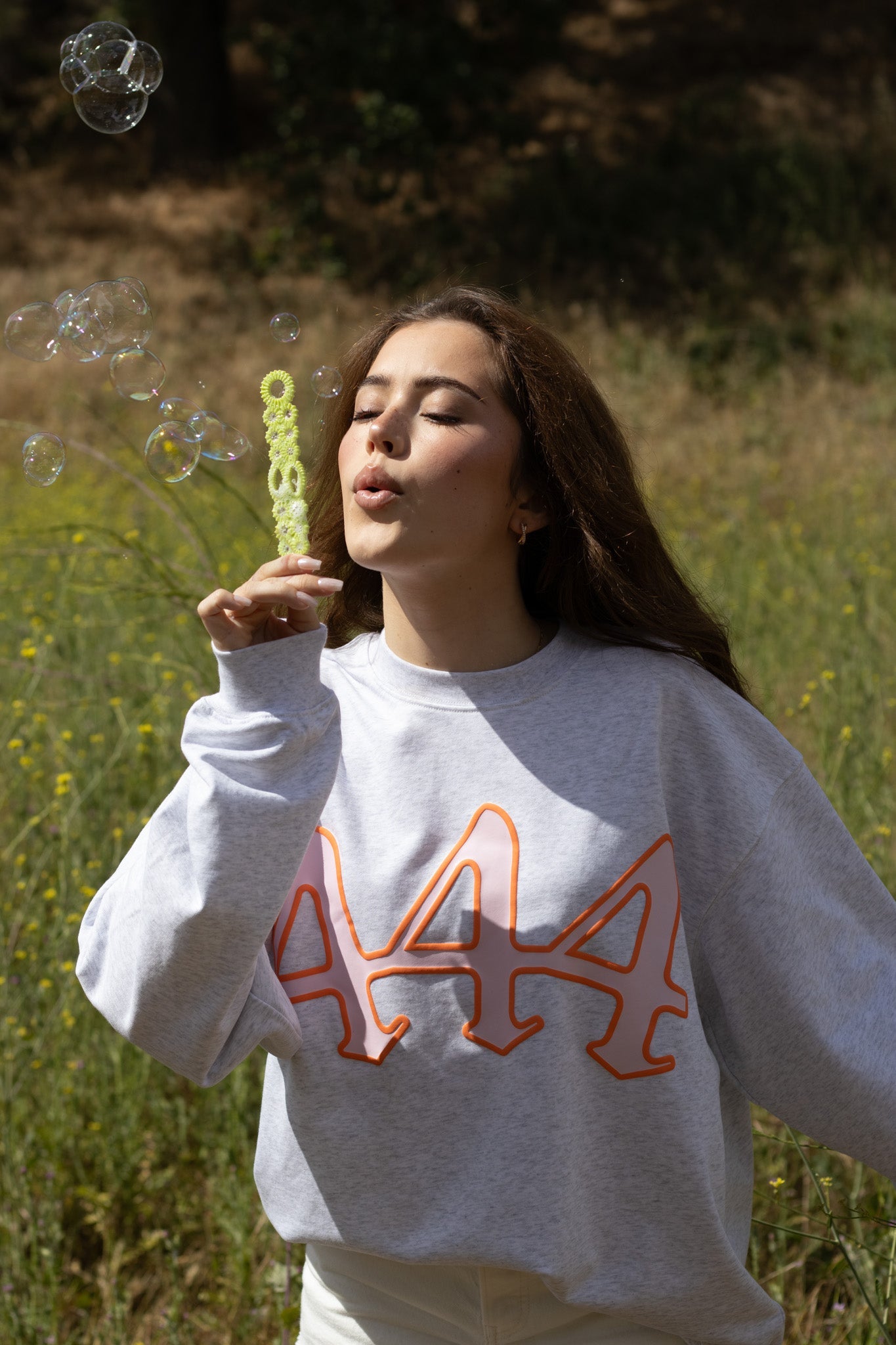 Woman blowing bubbles in a field wearing a white sweatshirt with orange text