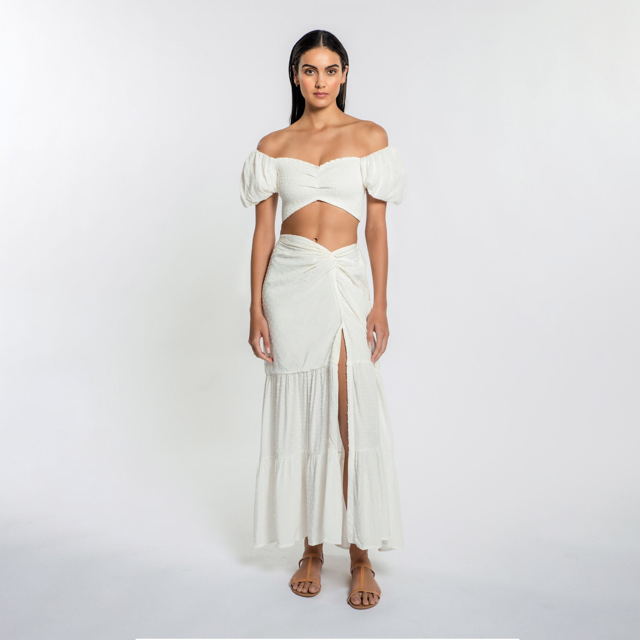 A woman wearing a white cotton skirt with an elastic waistband, thigh high front slit, and a knot detail at the waist. The skirt is lined and part of a matching set with a similar top.
